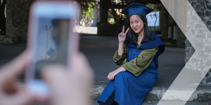Shot of a graduated college student smiling to the camera and making a V-sign after the graduation ceremony ends. She's sitting on the steps in her campus area while her unrecognizable friend takes her picture using a smartphone.