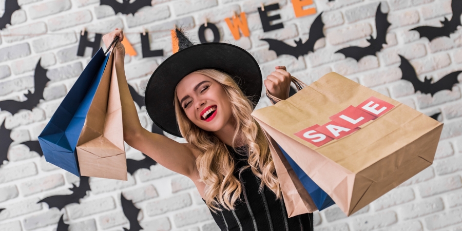 Woman in a witch hat holding shopping bags in front of a Halloween sign