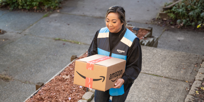 Woman in Amazon vest carrying Amazon box up a sidewalk for delivery