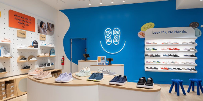 Image of the inside of Kizik's first physical retail location, shoes lined up on a table with a curving, modernist design in front of a blue wall with a while shelf built directly into it, above the shelf appear the words 
