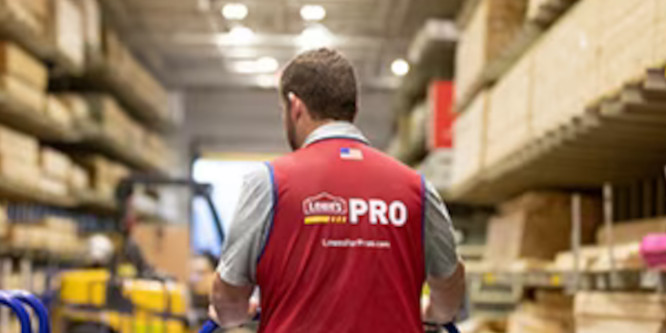 Lowe's employee pictured from the back wearing magenta smock that reads Lowe's Pro, in front of blurred Lowe's shelves/store interior