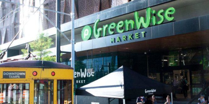 Picture of a GreenWise Market storefront. In front is an umbrella over a table where people are dining with the GreenWise Market logo on it. Next to them is a bus to downtown Tampa