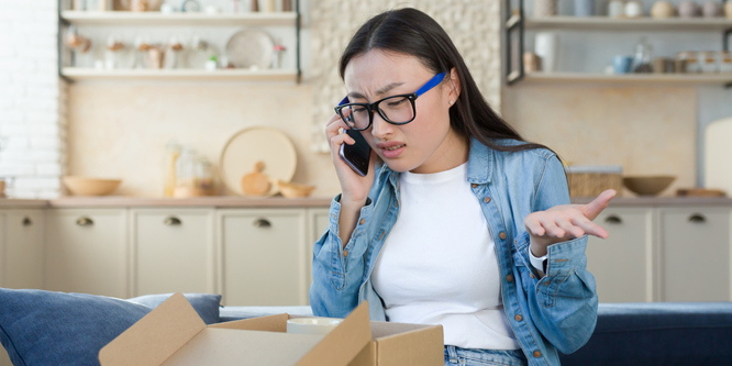 Unsatisfied woman received a broken parcel delivered from an online online store, Asian quarrels with the customer support service, talks to the operator on the phone, sitting home in the kitchen